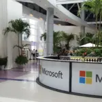 Trade show booth with a curved white counter and palms in a convention atrium