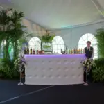 White tufted bar inside a draped tent surrounded by palms and tropical foliage