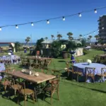 Beachfront lawn event under string lights with farm tables and blue-striped linens